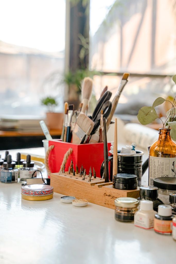 Collection of paint brushes of different sizes near jars of paints and ink bottles on white table with smooth surface in art studio with large window in sunlight